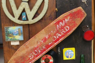 A license plate, small surf board, VW logo, picture of water and an old record on a wooden wall.